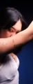 A person's face partly hidden by their arm, with tattoos on the forearm, wearing a white top and necklace. Dark background, soft light.