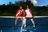 Two models in red and pink outfits by a tennis net on a blue court, with green trees and blue sky above.