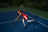 A person in a red and white tennis dress swings a racket on a blue court with a tennis ball nearby, bathed in sunlight.