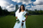 A model in a white dress and pearl necklace poses outdoors under a blue sky with clouds and bright sunlight.