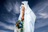 A model in a white draped garment holds a green and blue bouquet, brightly lit against a dark background.