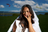 A model with long dark hair under dappled sunlight, wearing a white garment and pearl necklace, finger touching lips.