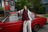 A person in a striped polo, grey hoodie, and sweatpants stands next to a red car. Green foliage in background.