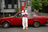 A model in a striped polo, grey hoodie, sweatpants, and tan boots leans on a red car by a red door.