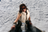 A model in sunglasses and a light fur vest, with dark hair, stands outdoors in bright sunlight against a snowy background.