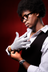 A model in black glasses, white shirt, black suspenders, and shiny studded arm wraps against a red background.
